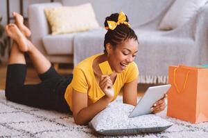Excited african girl showing credit card, using tablet, lying on the floor photo