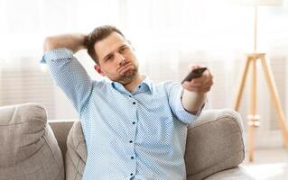 Nothing Interesting To Watch. Bored man in shirt watching TV on the couch at home and zapping, free space photo