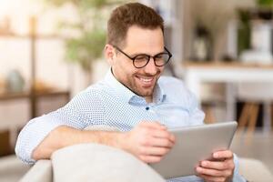 Reading News. Smiling man in spectacles using digital tablet sitting on the couch, copyspace photo