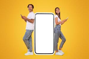 Modern Communication. Full Body Length Of Happy Cheerful Couple Leaning On Big Huge Smartphone With White Empty Blank Screen Using 2 Cell Phones, Chatting On Social Media Standing On Orange Background photo