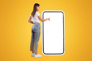 Full Body Length Back View Of Woman Using Big Smartphone With Blank White Screen And Touching Display Panel With Finger, Cheerful Lady Standing On Yellow Background, Ordering Food Delivery, Mock Up photo