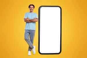 Full body length of smiling casual guy standing with folded arms near giant mobile phone, empty white screen template, orange studio background. Mock up for app or website, free space for advertising photo