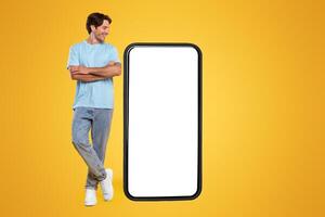 Cheerful smiling casual guy standing with crossed hands looking at giant big cell phone, empty white screen template, orange studio wall. Mock up for app or website, free copy space, full body length photo