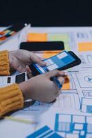 A person is using a smartphone on a table with a lot of paper and pens photo