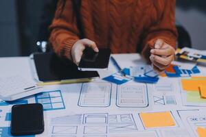 A person is using a cell phone and a laptop on a table with a lot of paper and photo