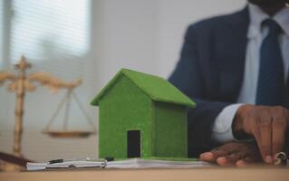 A man in a suit sits at a desk with a green house model photo