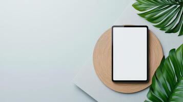 A tablet is on a wooden surface with green leaves in the background photo