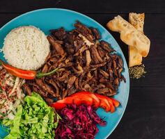 Delicious iskender kebab is served on a blue plate with rice, fresh vegetables and pita bread close up photo