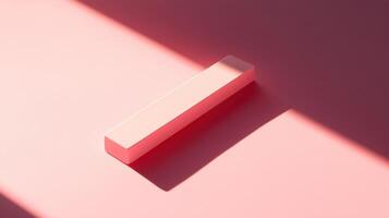 A single, light pink, rectangular prism sits on a pale pink surface, casting a shadow from a bright light source. photo