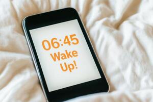 Smartphone alarm clock displaying 0645 wake up reminder on soft bed with gentle morning light photo