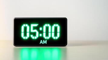 Sleek digital alarm clock displaying 05.00 AM in bright neon green light from a low angle on a smooth surface photo