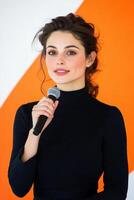 Engaging speaker holding microphone in front of vibrant orange backdrop during public event presentation photo