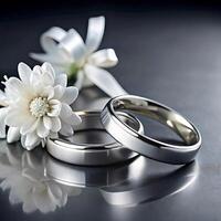 Two Elegant Wedding Rings Resting on a Dark Reflective Surface Near a Delicate White Flower photo