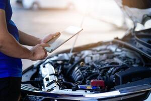 A man is looking at a tablet while standing next to a car. He is checking the engine of the car photo