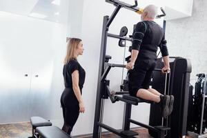 Elderly greyhaired man in ems suit is engaged on the uneven bars on the gravitron simulator with personal trainers in gym. photo