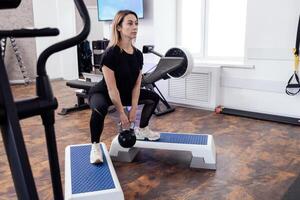 Sportswoman in black sportswear standing on step platforms and doing squats with kettlebell. photo