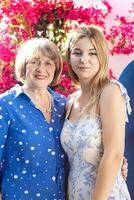 Portrait of a beautiful girl and her grandmother on the background of a crimson flowering tree. A smiling pensioner and her sweet granddaughter pose outdoors. photo