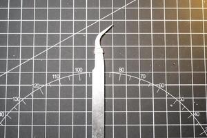 Metal precision tweezer with pointed tip. Tweezer is on a black cutting mat with lines. A tool that functions to clamp and pull out small objects. Negative Space. Beauty and grooming tools photo