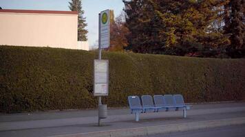 Temporary bus stop in Bavaria, Germany, during autumn season. It features blue benches, green hedge, street sign with timetable and autumn trees, with no roof, creating peaceful suburban atmosphere. video