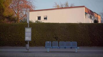 This bus stop in Bavaria, Germany, is set in autumn. It has blue benches, a green hedge, a timetable sign, and autumn trees, with no roof and no people, creating a peaceful suburban atmosphere video
