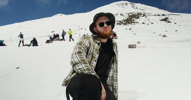 A man with a beard and sunglasses sitting on a snow covered mountain photo