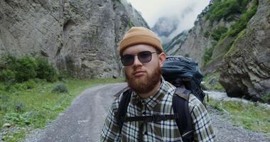 The Caucasus. A young fashionably dressed man with a beard, with a tourist backpack, stands on the road among the rocks and smiles while looking at the camera. Mountain peaks are covered with clouds photo