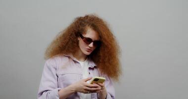 A beautiful red-haired girl with lush hair in sunglasses uses the phone, texting and laughing. Shooting against a light background. photo