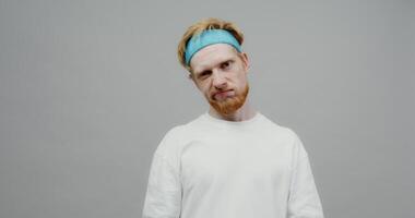 A red-haired guy with a beard and a sports band on his head, looking bored, trains his biceps with small dumbbells. Shooting on a light solid background. photo