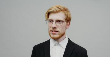 A red-haired guy with a beard in glasses, dressed in a business black suit, is seriously looking at the camera, telling something. Shooting on a light solid background. photo