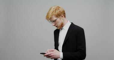 A serious red-haired guy with a beard in glasses, dressed in a business black suit, is texting on a mobile phone. Shooting on a light solid background. photo