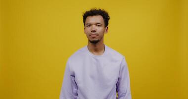 Young mulatto man with black curly hair waves his hand hello and smiles while looking at the camera. Shooting against a solid yellow background. photo