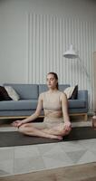 A beautiful girl of European appearance with gathered hair without makeup, dressed in sportswear, sits in a lotus position with closed eyes on a gymnastics rug, in a modern interior, vertical photo