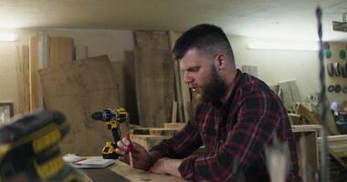 a European man with a beard and dark hair in a plaid shirt talking by call on a laptop, actively explaining something gesticulating with hands and smiles in a carpentry workshop photo