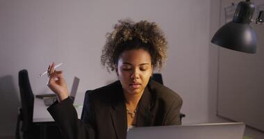 dark-skinned girl in black business jacket without shirt with nose ring chain, around her neck twists pen in her hand looks at laptop screen makes notes in notebook in office under artificial lighting photo