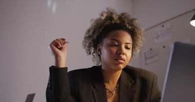 dark-skinned girl in black business jacket without shirt, with nose ring and chain around her neck twists a pen in her hand typing and looking at a laptop screen in an office under artificial lighting photo