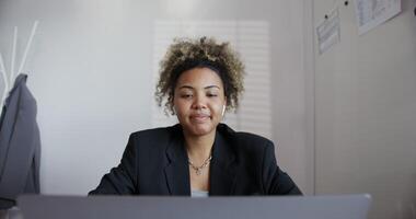 beautiful black-skinned girl with afro hairstyle in black business jacket, with earring in the nose and chain on her neck is greets and actively talking on open laptop and headphones in ears close-up photo