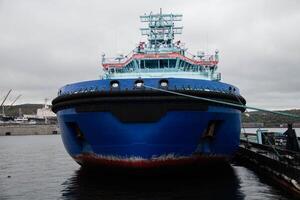 Large Blue Tugboat Docked at busy Shipyard under overcast Cloudy Sky. Visible Maritime infrastructure and Industrial background add context to Vessel's functional role in Port operations photo