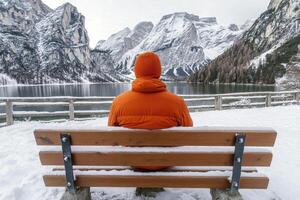 Person in Orange Jacket on Snowy Bench Overlooking Winter Lake and Mountains photo