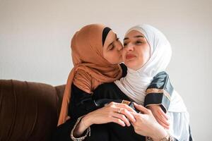 Happy Muslim family at home young mother and child daughter in hijab kiss and hug with love on sofa smiling and playing together for bonding with affection on mothers day. photo
