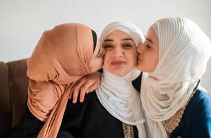 Happy Muslim family at home young mother and two child daughters in hijab kiss and hug with love on sofa smiling and playing together for bonding with affection on mothers day. photo