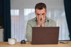 Tired man working overtime, sitting at the desk in front of a laptop and rubbing eyes with hands. photo