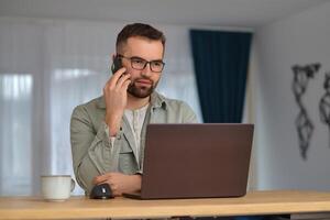 A young man talking on his phone and using a laptop while working at home office. photo