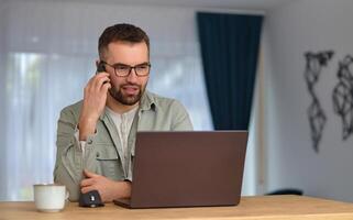 A happy man talking on his phone and using a desktop computer while working at home office. photo