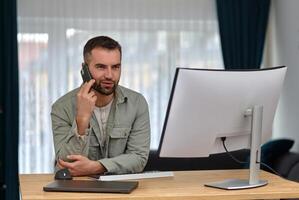 A happy man talking on his phone and using a desktop computer while working at home. photo