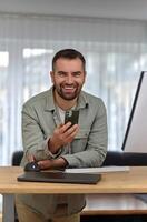 A young happy man using his phone mobile while working at home office, looking at the camera and smiling. photo
