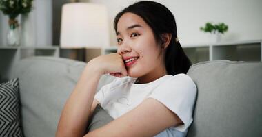 Handheld shot, Young Asian woman sits on a sofa relax and smiles happily after hearing something that makes her feel good or relieved. video