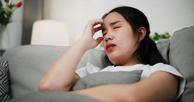 Handheld shot, Asian woman sitting alone on the sofa, looking sad, hold the head with hand while thinking about something in the living room at home. Facial expression concept. video
