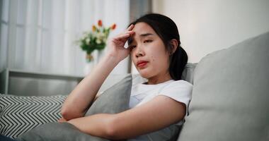 Handheld low angle view shot, Asian woman sitting alone on the sofa, looking sad, hold the head with hand while thinking about something in the living room at home. Facial expression concept. video