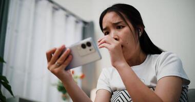 Handheld low angle view shot, Young asian woman wearing wireless headphones sits on a sofa with eyes wide open and mouth slightly open, indicating shock or surprise while looks at mobile phone. video
