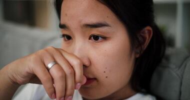 Handheld selective focus shot, Young Asian woman sits on a sofa with hand touch lips while thinking about something or feeling confused and hesitant. Facial expression concept video